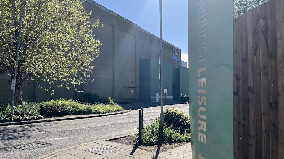 A side entrance to Cambridge Leisure Park, showing the side of the big brick building housing the cinema. There is greenery and a road. It is a sunny evening and there is some lens flare on the image.