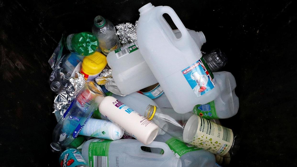 A collection of plastic milk bottles, metal containers and tin foil in a heap against a black background. The pile also includes a shampoo and drinks bottles. 
